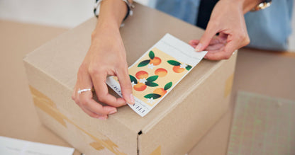 Impression M rectangular sticker with orange fruit pattern being applied to a cardboard shipping box